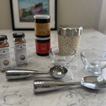 A tabletop arrangement of baking and dessert-making items on a marble surface, including two jars of Watkins sprinkles, two stacked jars of Nutella chocolate spread, a patterned metal cup, two clear glass bowls, and two metal ice cream scoops.