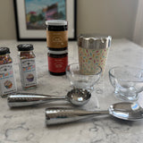 A tabletop arrangement of baking and dessert-making items on a marble surface, including two jars of Watkins sprinkles, two stacked jars of Nutella chocolate spread, a patterned metal cup, two clear glass bowls, and two metal ice cream scoops.