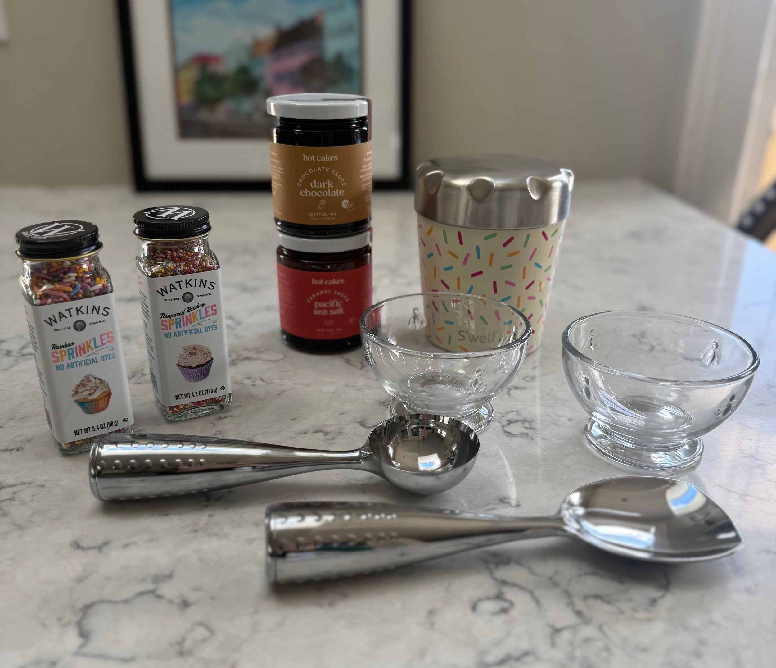 A tabletop arrangement of baking and dessert-making items on a marble surface, including two jars of Watkins sprinkles, two stacked jars of Nutella chocolate spread, a patterned metal cup, two clear glass bowls, and two metal ice cream scoops.